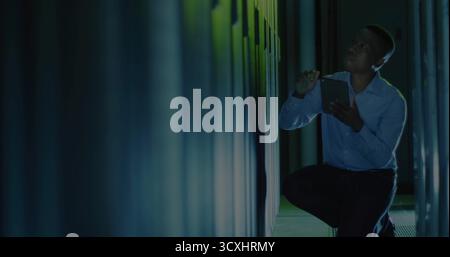 Kneeling IT technician inspecting servers in data center corridor, with tablet, stylus, copy space Stock Photo