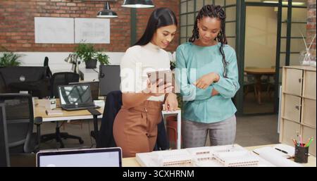 Colleagues wearing office attire using tablet while reviewing scale model at desk, with laptops Stock Photo