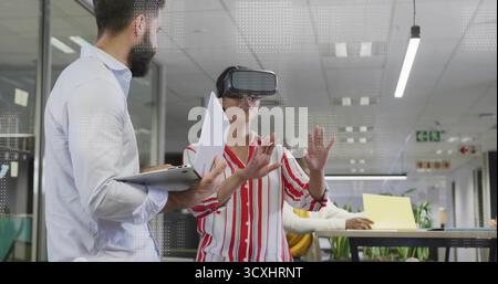 Gesturing woman wearing VR headset in open-plan office with laptops, paper and bar-height table Stock Photo
