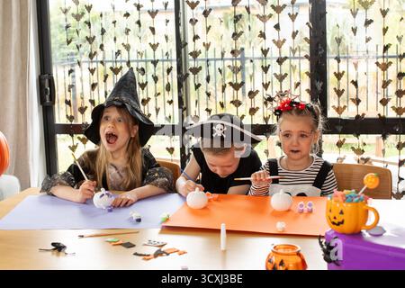 Happy children paint pumpkins in Halloween costumes for a festive craft activity. Craft workshop.  Stock Photo
