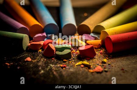 Colorful chalk sticks arranged in a semi-circle on a dark textured surface, with broken pieces scattered in the center. Stock Photo
