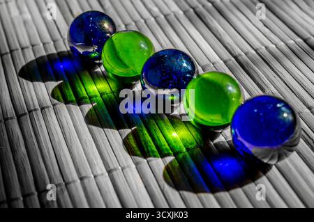 Five glass marbles in alternating blue and green hues placed on a bamboo mat, casting vivid colored shadows due to strong directional light. Stock Photo