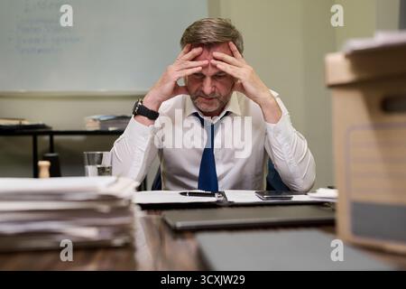 Middle aged Caucasian man sitting at desk holding head in hands appearing stressed, while working with documents in office environment surrounded by paperwork and folders Stock Photo