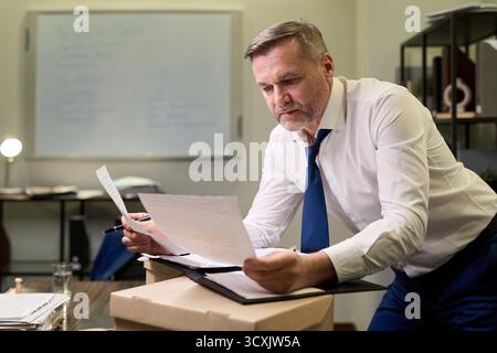 Middle aged Caucasian man reviewing documents while leaning over desk in office, holding paperwork and pen, appearing focused on reading and analyzing information during work Stock Photo