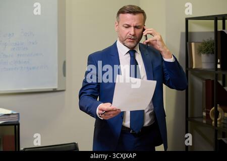 Middle aged Caucasian man standing in office talking on smartphone while holding document, appearing focused and engaged in business conversation near whiteboard and shelves Stock Photo