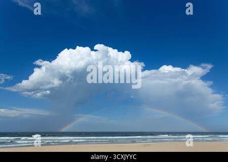 Rainbow forming under the storm clouds over the ocean against a blue sky. Stock Photo