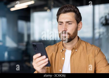Bearded man intently focused on his smartphone, networking and managing business apps in a modern coworking space, checking messages, browsing data an Stock Photo
