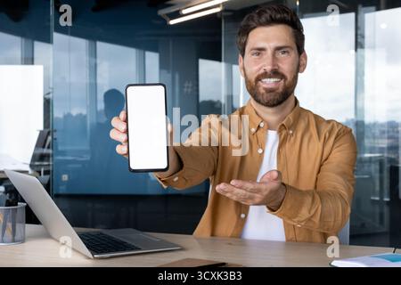 Smiling businessman holding a modern smartphone with a blank white screen for mockup, gesturing while presenting new app or service in a contemporary Stock Photo