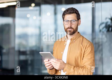 Confident bearded man with glasses standing in a modern office, holding a tablet and engaging with digital technology for work, communication and busi Stock Photo
