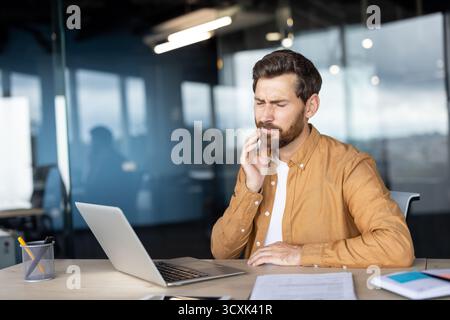 Young man working on laptop in a modern office, experiencing severe toothache and jaw pain, touching his cheek with hand, showing discomfort and denta Stock Photo