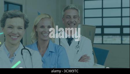 Posing healthcare team wearing white lab coats with stethoscopes and blue scrubs in clinical office Stock Photo