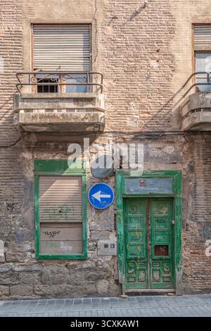 Weathered facade of old building with green wooden door, concrete balconies and exposed brick showing urban decay. Authentic Spanish architecture for Stock Photo