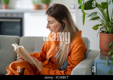 Middle-aged woman deeply absorbing story in paper book seated comfortably sideways on home sofa Stock Photo
