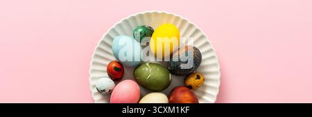 A collection of vibrant Easter eggs in various colors is artfully arranged on a white plate, set against a soft pink background, capturing the essence of the holiday's decorations, banner Stock Photo