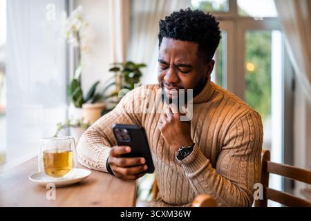 A young black man sits at the bar, using smartphone, messaging online. He is anxious and distraught. Stock Photo