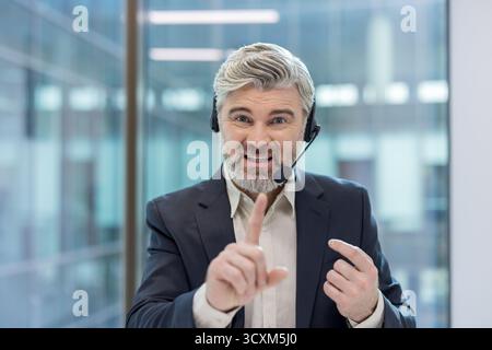 Mature man with a beard and headset talking, actively communicating with customers, providing support, and gesticulating with his finger in a modern o Stock Photo