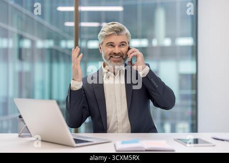 Senior businessman standing at office desk, talking on a mobile phone, smiling cheerfully and gesturing with his hand, interacting in a corporate work Stock Photo