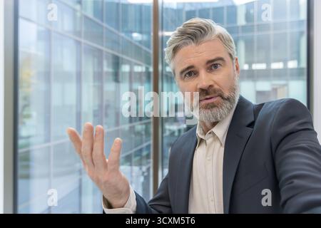 Middle-aged businessman communicating and gesturing during a video call or online meeting, creating an engaging presentation from a contemporary offic Stock Photo