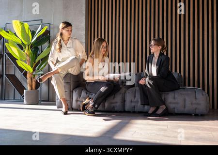 Three businesswomen are sitting on a modern office sofa, discussing important partnership details and collaborating on paperwork, representing teamwor Stock Photo