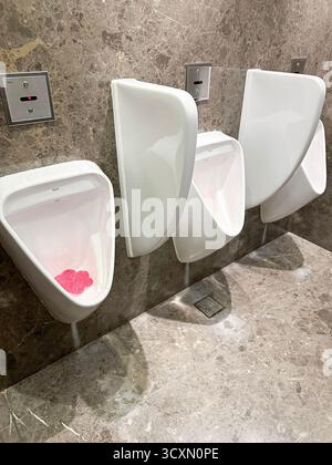 Three white porcelain urinals with a pink urinal cake on a marble tiled bathroom wall in a public restroom Stock Photo