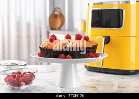Freshly baked muffins with raspberries and air fryer on white marble table in kitchen, closeup Stock Photo