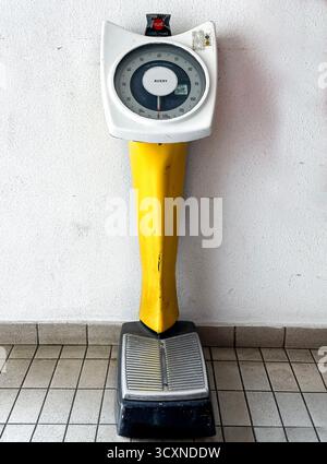 Vintage Avery coin operated weighing scales with a yellow base against a white wall, standing on a tiled floor in public toilet kuala lumpur malaysia Stock Photo
