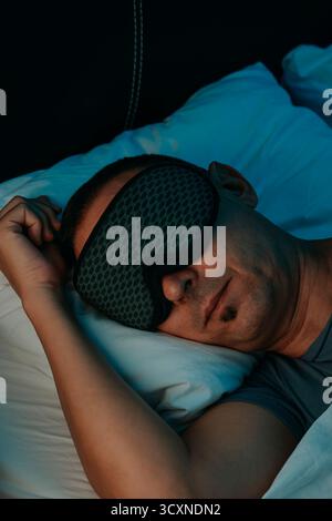 closeup of a man in a bedroom wearing a patterned sleep mask, his face resting on a pillow while he lies under a bed sheet with one hand near his head Stock Photo