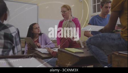 Collaborating students pointing at tablet screen in classroom, passing papers at wooden desks Stock Photo