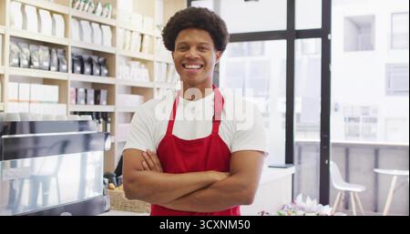 Smiling barista in red apron folding arms behind counter at coffee shop, with espresso machine Stock Photo