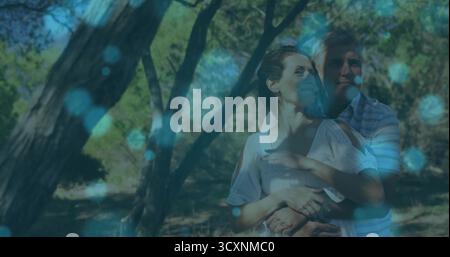 Embracing middle-aged couple standing among sunlit woodland trunks, with ethereal blue light orbs Stock Photo