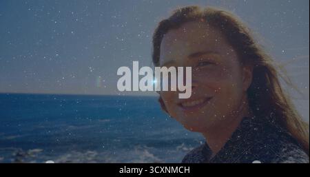 Standing woman in geometric shirt smiling at camera on beach edge, sunlight reflecting off water Stock Photo