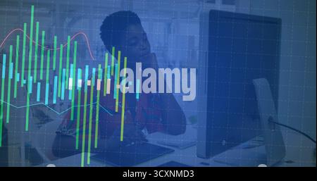 Gazing office worker wearing red blouse resting chin on hand at desk with computer monitor, charts Stock Photo