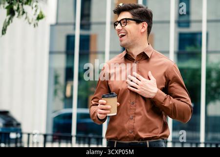 Young professional man in brown shirt holding coffee, smiling outdoors in urban city environment, showcasing confidence Stock Photo