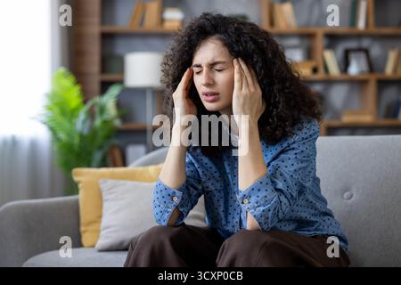 Young woman feeling intense pain and pressure in her head, closing eyes and pressing hands on her temples, sitting on a sofa at home experiencing disc Stock Photo