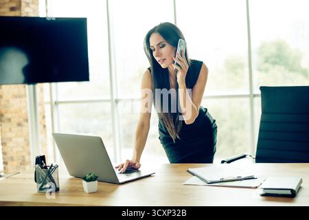 Confident businesswoman in a modern office multitasking on a call and laptop, showcasing efficiency and poise Stock Photo