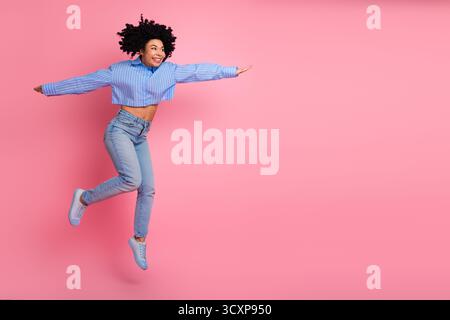 Happy young woman leaping energetically with joyful expression in a colorful outfit on a pink background, feeling inspired Stock Photo