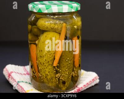 Homemade pickled cucumbers and carrots in a glass jar, spiced with mustard seeds, resting on a checkered kitchen towel against a dark background Stock Photo