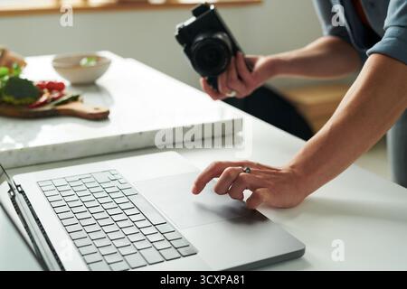 Man Using Laptop While Holding Camera in Kitchen Setting Stock Photo