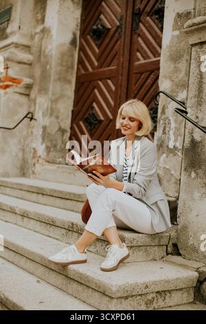 A young woman with short blonde hair sits on the steps of an old building. She reads a book while dressed in a blazer and casual shoes, enjoying a sun Stock Photo