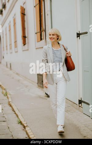 A fashionable woman in a light gray blazer and striped shirt strolls along a narrow street lined with charming buildings, enjoying a sunny day. Stock Photo