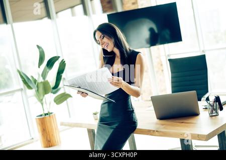 Confident businesswoman reviewing documents in modern office setting while discussing plans on the phone Stock Photo