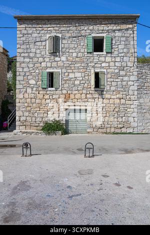 A rustic stone building with green wooden shutters sits on a quiet street under a clear blue sky. Weathered walls, small plants, and simple metal bike Stock Photo