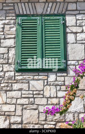 A rustic stone house with weathered green shutters sits beside vibrant bougainvillea in full bloom under a clear blue sky. Charming, warm, and picture Stock Photo