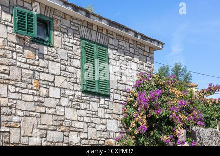 A rustic stone house with weathered green shutters sits beside vibrant bougainvillea in full bloom under a clear blue sky. Charming, warm, and picture Stock Photo