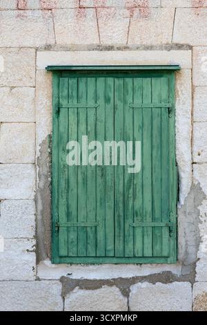 A weathered stone wall frames a faded red inscription above a worn green wooden shutter. The rustic facade evokes heritage, aging textures, and quiet, Stock Photo