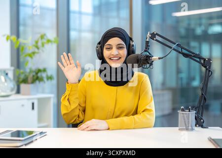 Muslim woman wearing a hijab and headphones, working as a radio host or podcaster in a modern studio, happily greeting an audience while recording con Stock Photo