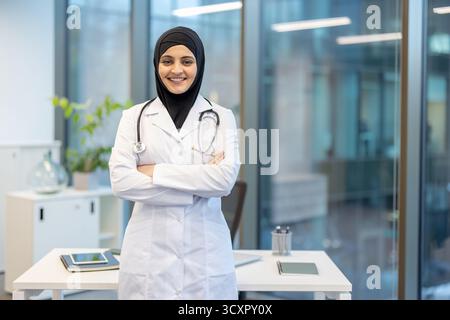 Muslim woman doctor wearing a hijab and lab coat with a stethoscope around her neck, standing with arms crossed and smiling confidently in a modern me Stock Photo