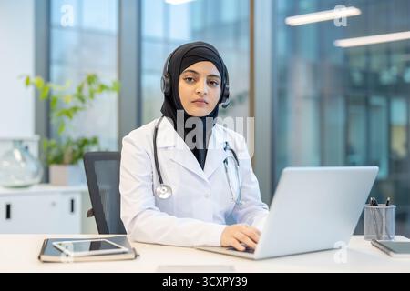 Muslim female doctor wearing a hijab and stethoscope, smiling while sitting at a desk and using a headset and laptop for a remote online medical consu Stock Photo