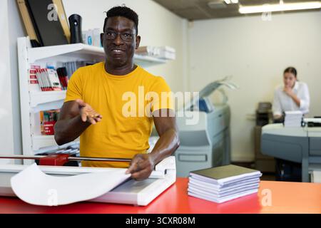 Portrait of happy middle-aged African American man using paper cutter during work in the printer shop Stock Photo