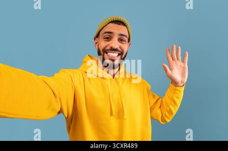 Hello. Happy arab guy taking selfie and waving hand at camera, gesturing hi while making photo over blue background Stock Photo